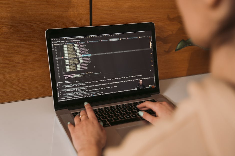A person with light skin and short hair is sitting at a white table, working on a laptop that displays lines of code on its screen. The laptop is placed in front of a wooden backdrop, suggesting an in