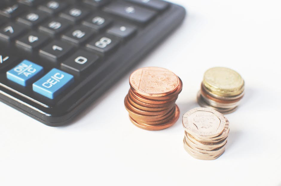 Close-up image showing a black calculator with blue and white buttons beside three separate stacks of British coins, including copper, silver, and gold-colored coins, arranged on a white surface. The 